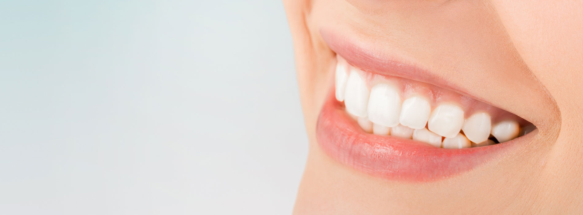 The image shows a close-up of a smiling person s teeth, with their lips slightly parted, set against a blurred background that appears to be a gradient of colors from light at the top to darker tones near the bottom.