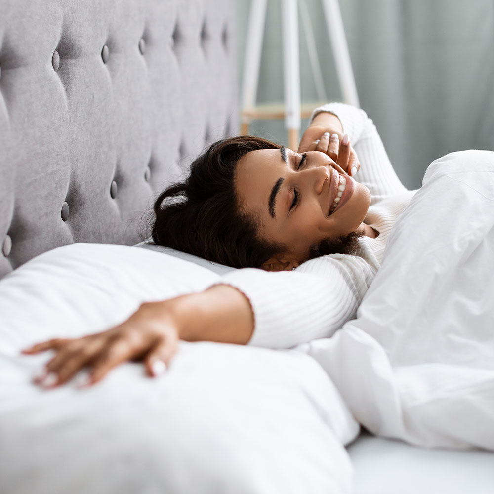 A person lying on their back with arms crossed, smiling contentedly while resting on a bed with white sheets.
