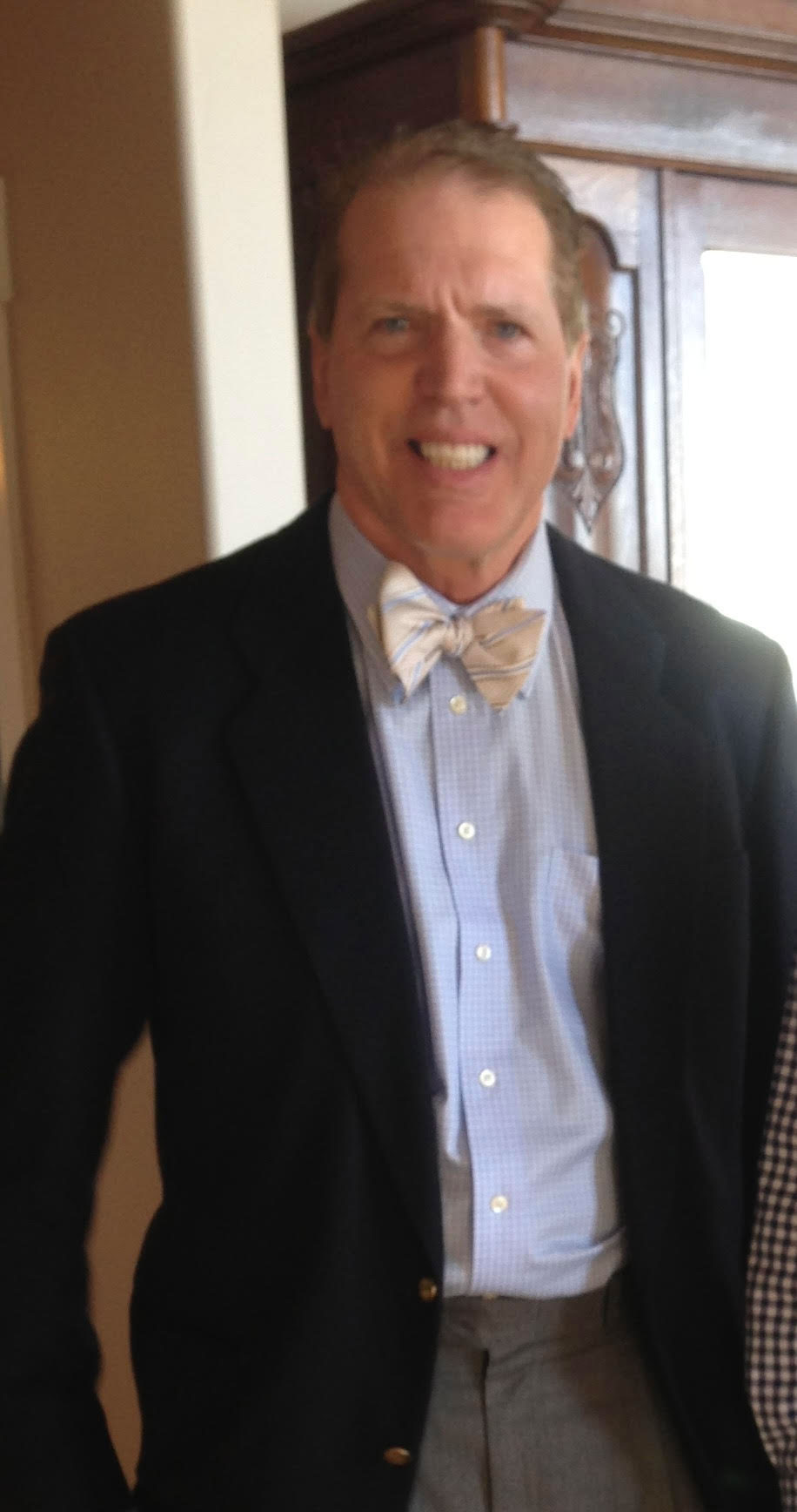The image features a man wearing a suit with a bow tie, standing indoors, smiling at the camera.