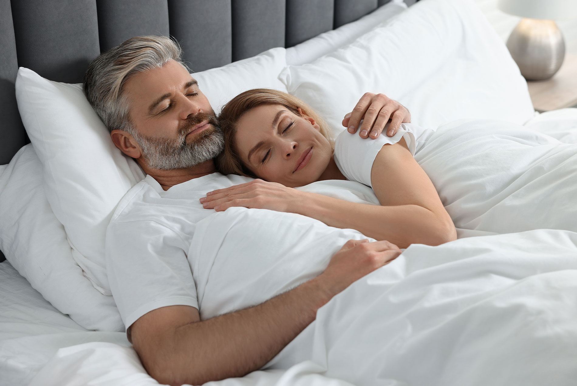 A man and woman are lying in bed, embracing each other with their eyes closed, suggesting a moment of intimacy or rest.